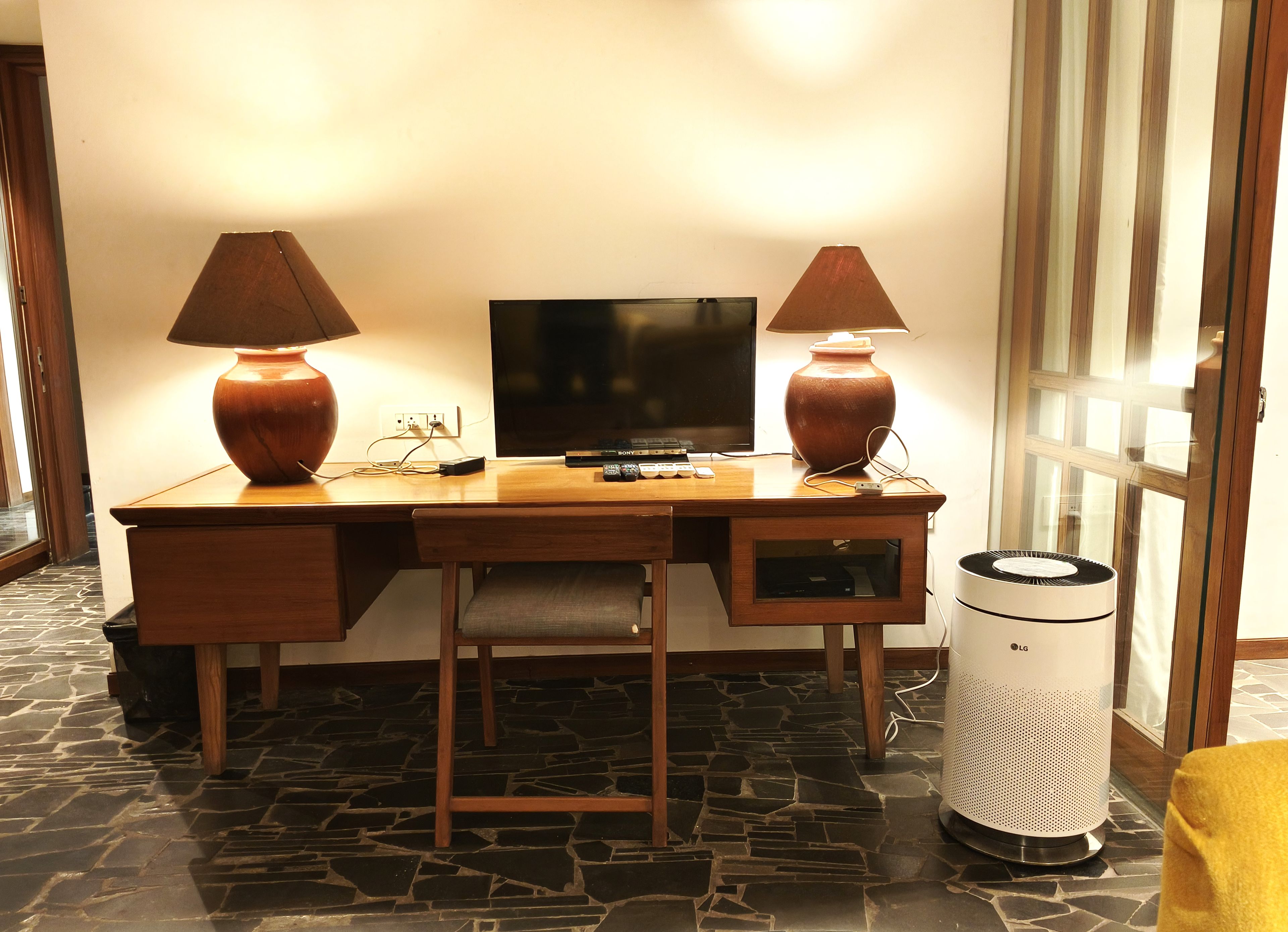 Un escritorio de madera con dos lámparas, un televisor y un purificador de aire LG en una habitación con suelo de piedra y una puerta de cristal.