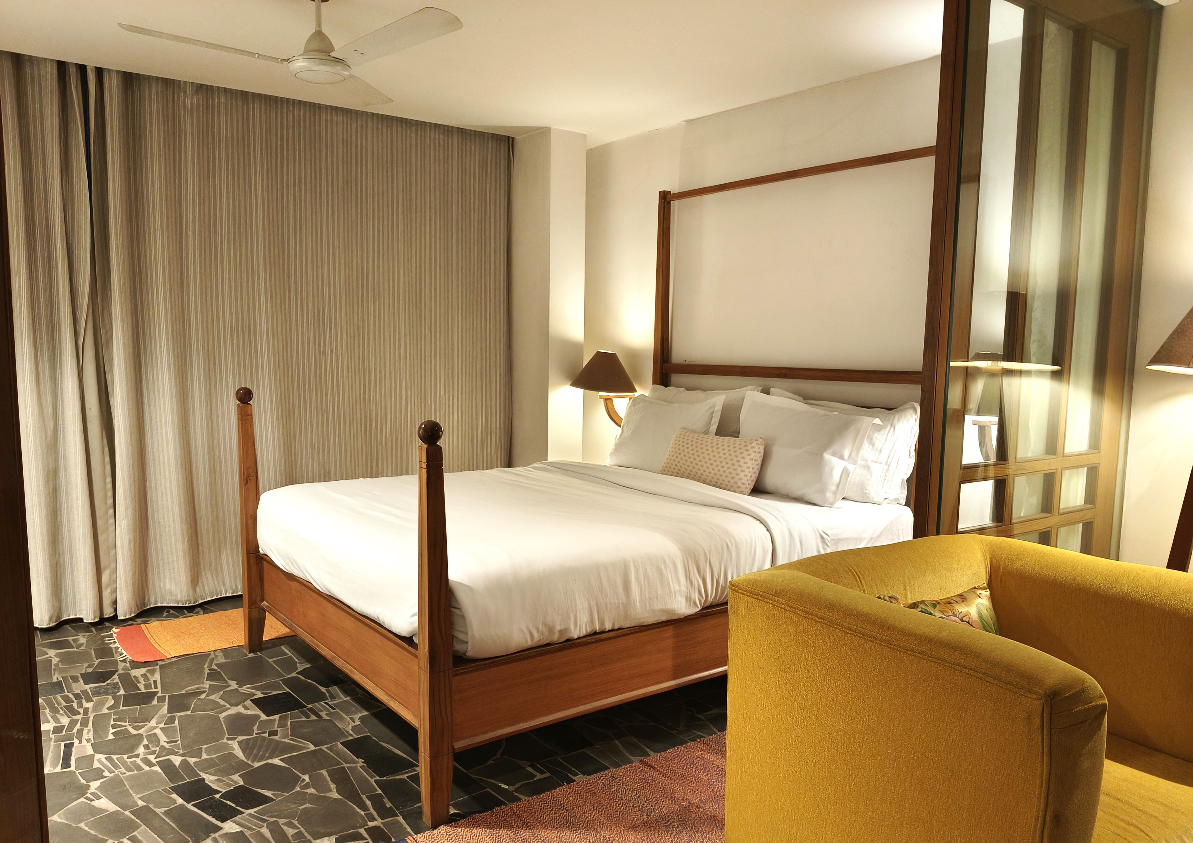 A hotel room with a four-poster bed, a wooden headboard, a yellow armchair, and a large window with curtains.