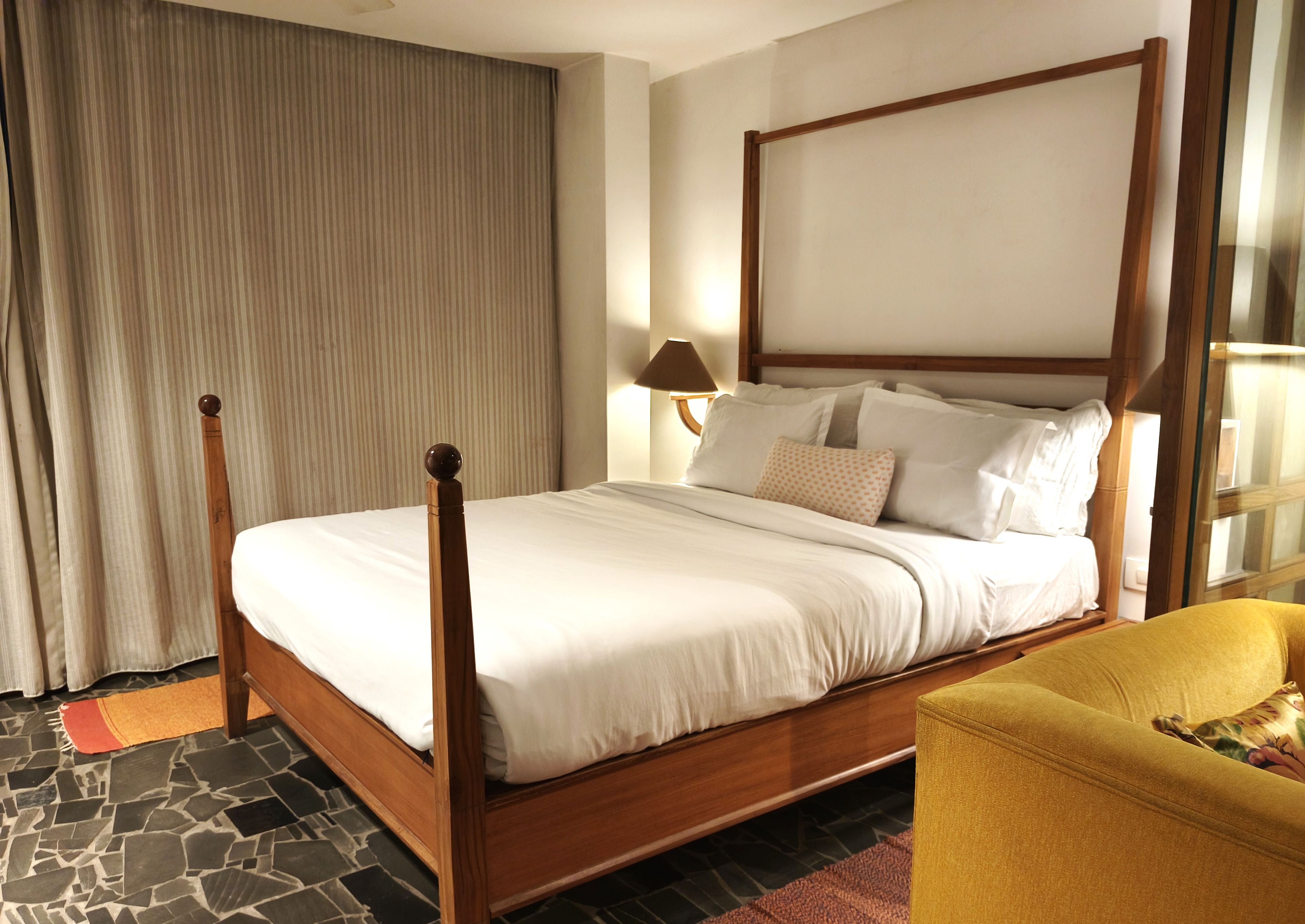 A four-poster bed with a wooden frame and white bedding, a stone-patterned floor, a yellow armchair, and a large window with curtains.