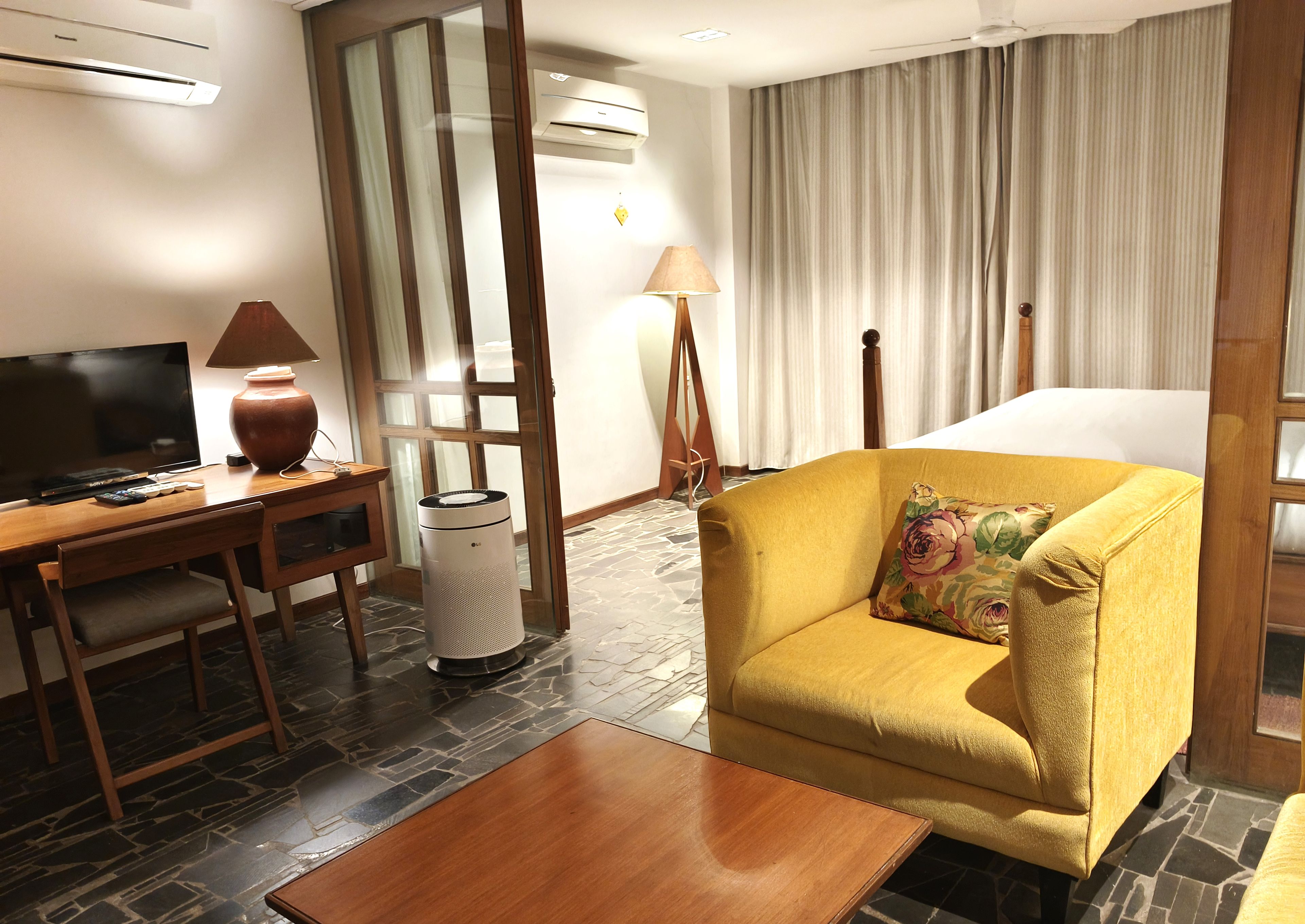 A hotel room with a wooden desk, a television, a sofa with a floral pillow, a standing lamp, and a bed with curtains.