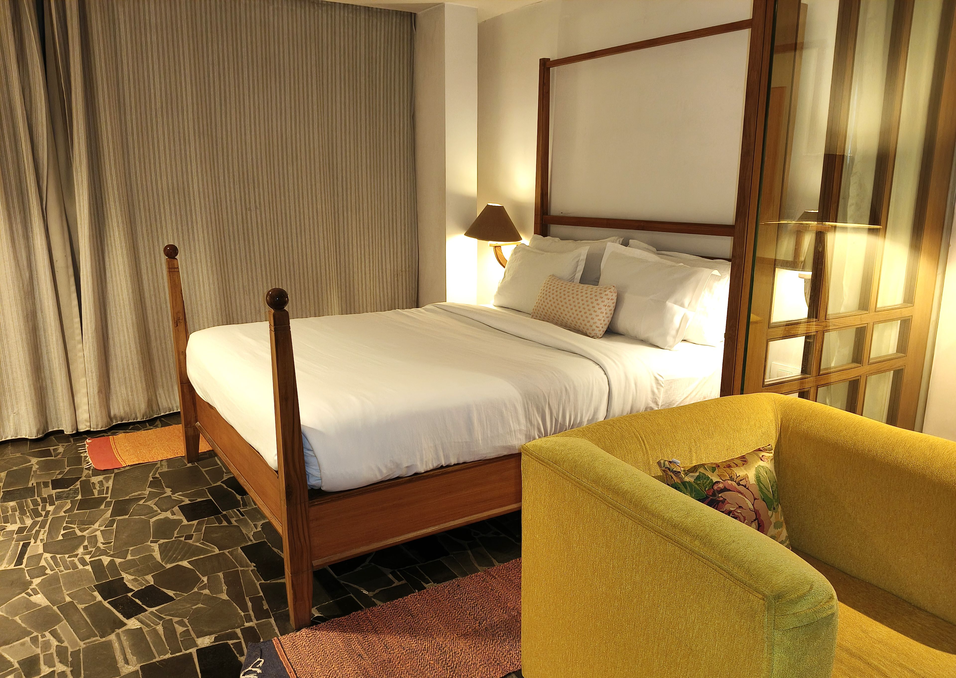 A neatly made bed with a wooden headboard, a yellow armchair, and a patterned rug on a stone floor.