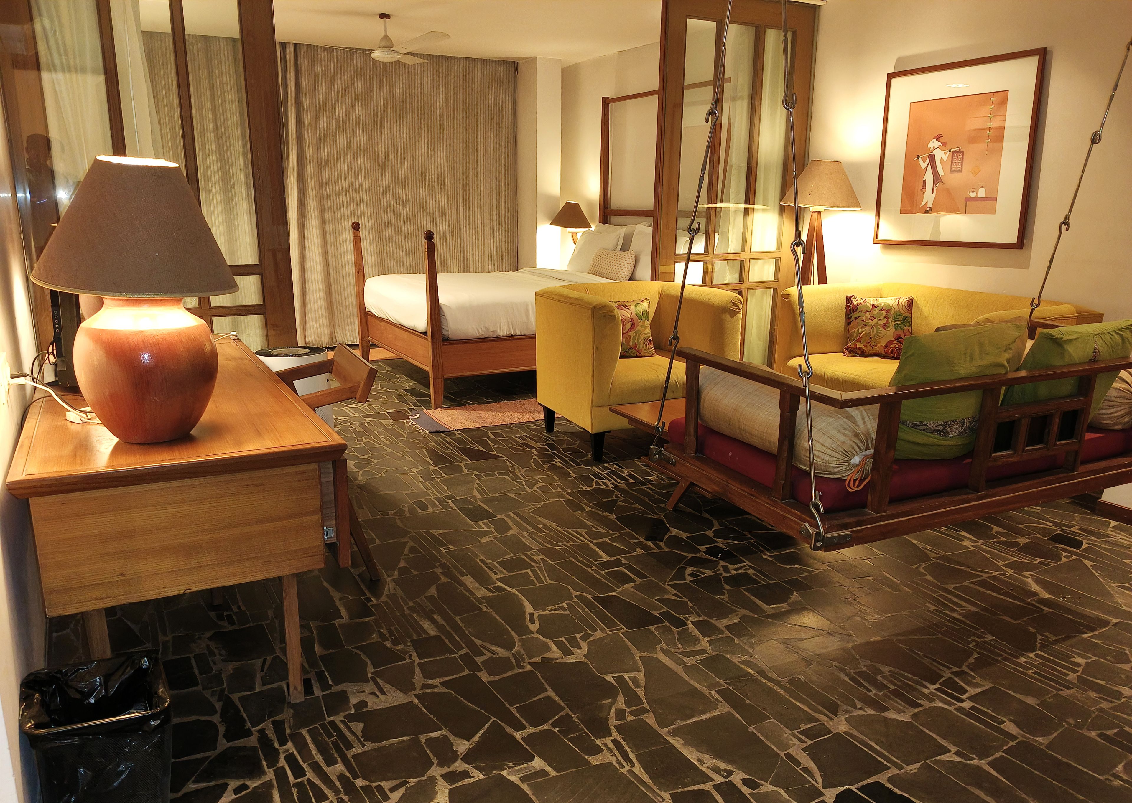 A hotel room with a bed, a sofa, a wooden table with a lamp, a hanging chair, and a stone-patterned floor.
