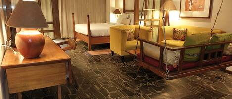 A hotel room with a bed, a sofa, a wooden table with a lamp, a hanging chair, and a stone-patterned floor.