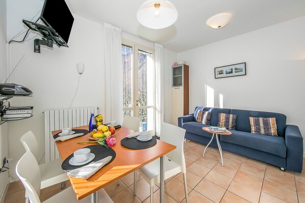 A modern living room with a blue sofa, a dining table with a fruit bowl, and a TV mounted on the wall.