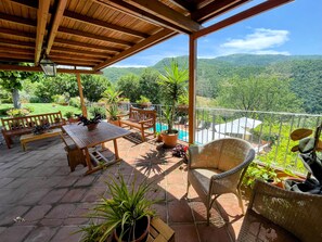 A covered patio with a wooden bench, table, and wicker chair, surrounded by potted plants and overlooking a lush valley.
