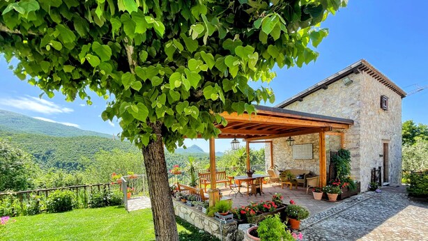 Ein Steinhaus mit einer überdachten Terrasse, Gartenmöbeln und Blumentöpfen.