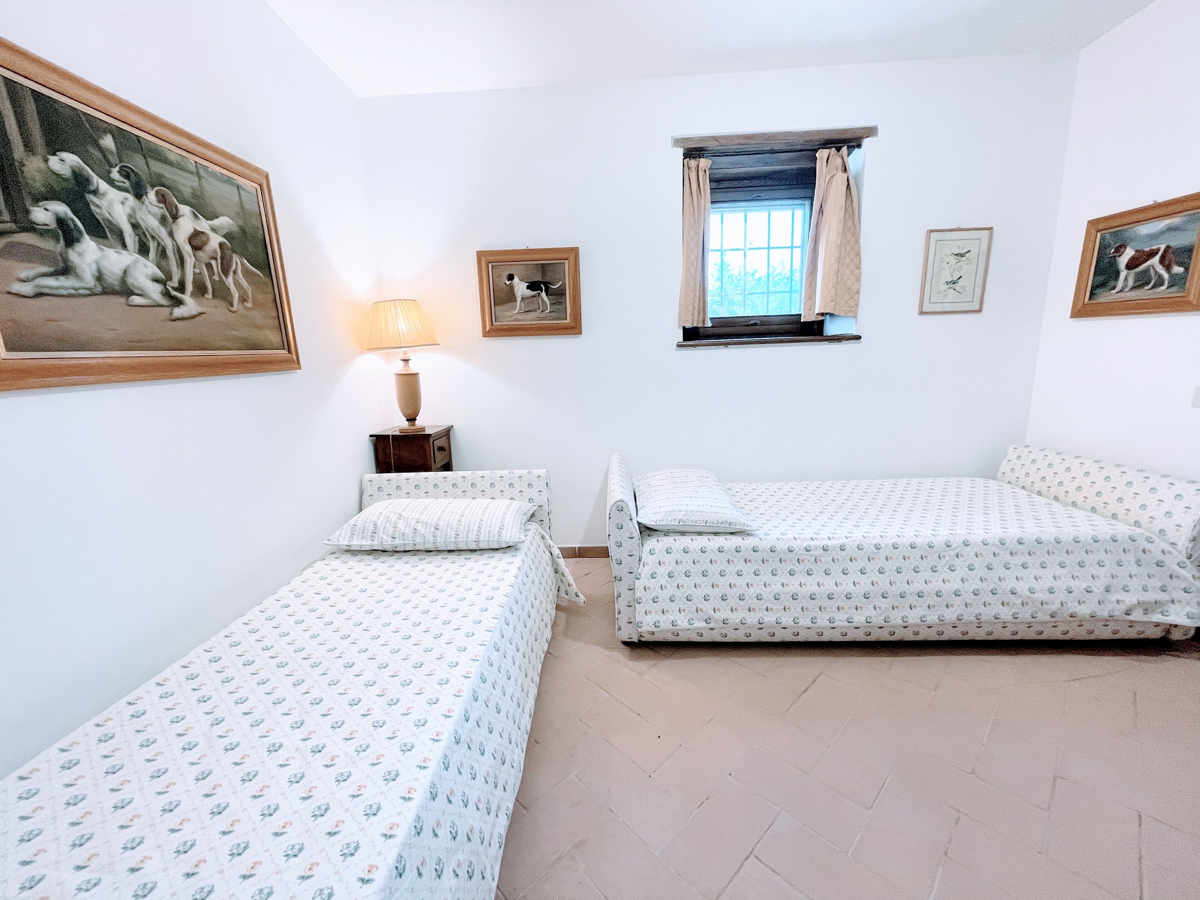 Una habitación con dos camas, una ventana con cortinas, una lámpara y cuadros en las paredes.