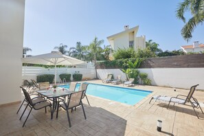A pool area with lounge chairs, a table with chairs, and a white umbrella. - Protaras Holiday Villa Dv27 (Protaras)