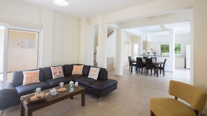 A modern living room with a dark sofa, coffee table, and yellow armchair. A dining area with a table and chairs is visible in the background. - Protaras Delfini Villa Dv4 (Protaras)