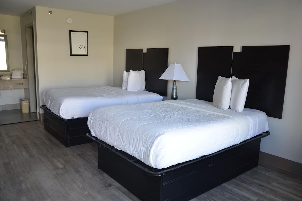 Quarto de hotel com duas camas, uma mesinha de cabeceira com abajur, um quadro na parede e um espelho sobre a porta.