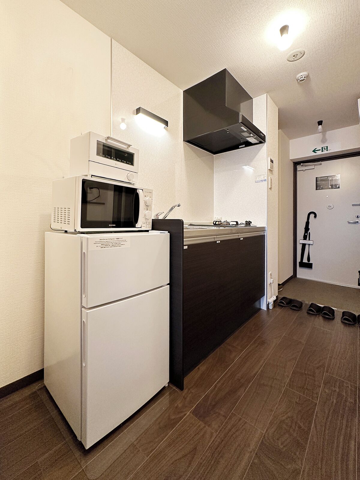 A compact kitchen with a microwave, refrigerator, and stove. There is a black hood above the stove, and a door with an exit sign. Shoes are placed on the floor near the door.