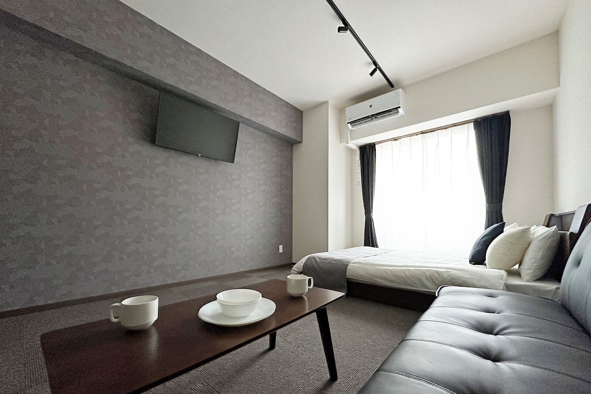 A modern hotel room with a sofa, a small table with cups, a bed, and a TV mounted on the wall.