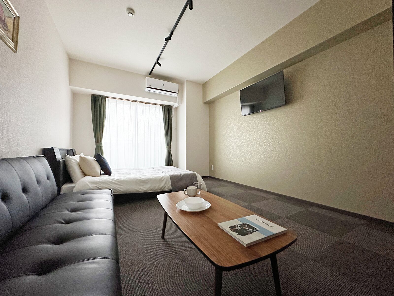 A hotel room with a sofa, a bed, a coffee table with a book, an air conditioning unit, and a TV mounted on the wall.
