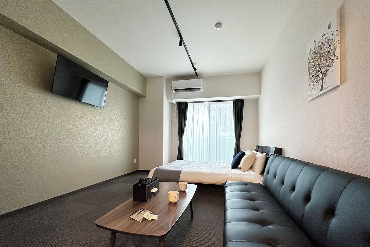 A hotel room with a bed, a sofa, a coffee table, a TV mounted on the wall, and an air conditioning unit.