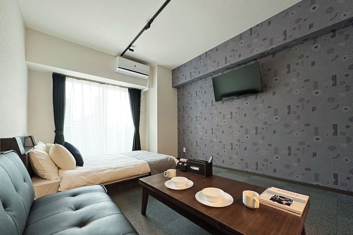 A modern hotel room with a bed, a sofa, a dining table with plates and cups, a flat-screen TV, and an air conditioning unit.