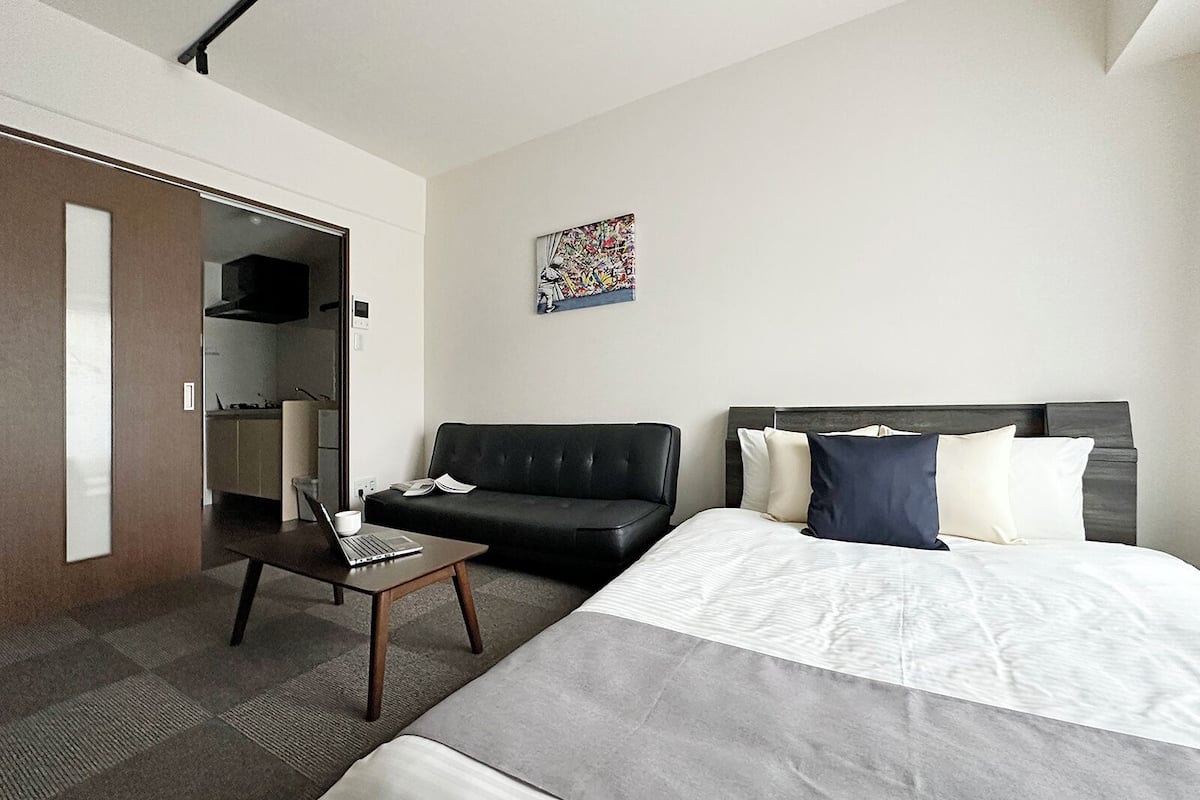 A modern hotel room with a large bed, a small sofa, a desk with a laptop, and a kitchen area visible through an open door.