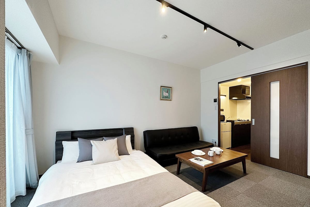 A hotel room with a large bed, a sofa, a small table with cups and a book, and a kitchenette visible through an open door.