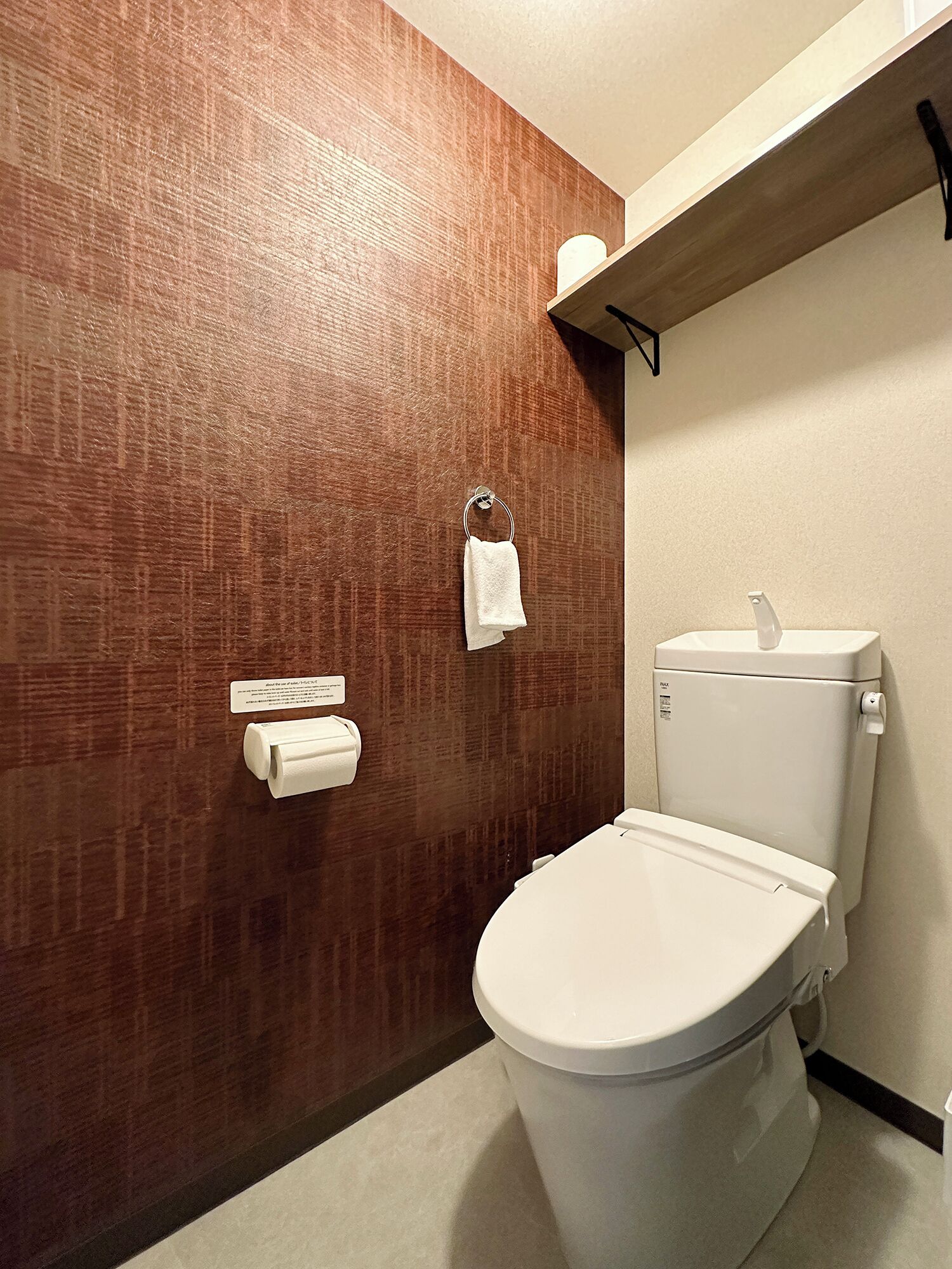 Uma casa de banho com parede texturizada em madeira, uma sanita, um dispensador de papel de cozinha e uma prateleira com rolo de papel higiénico.