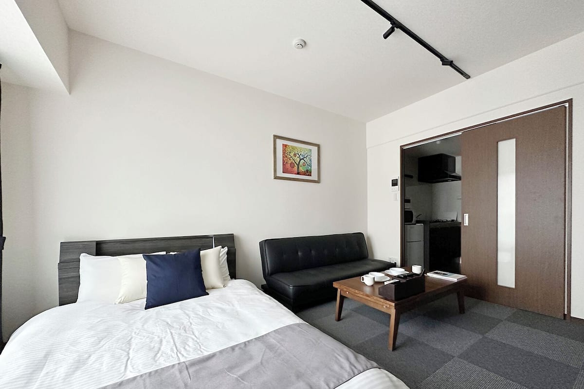 A hotel room with a bed, a black leather sofa, a small wooden table with cups and a book, and a kitchenette in the background.
