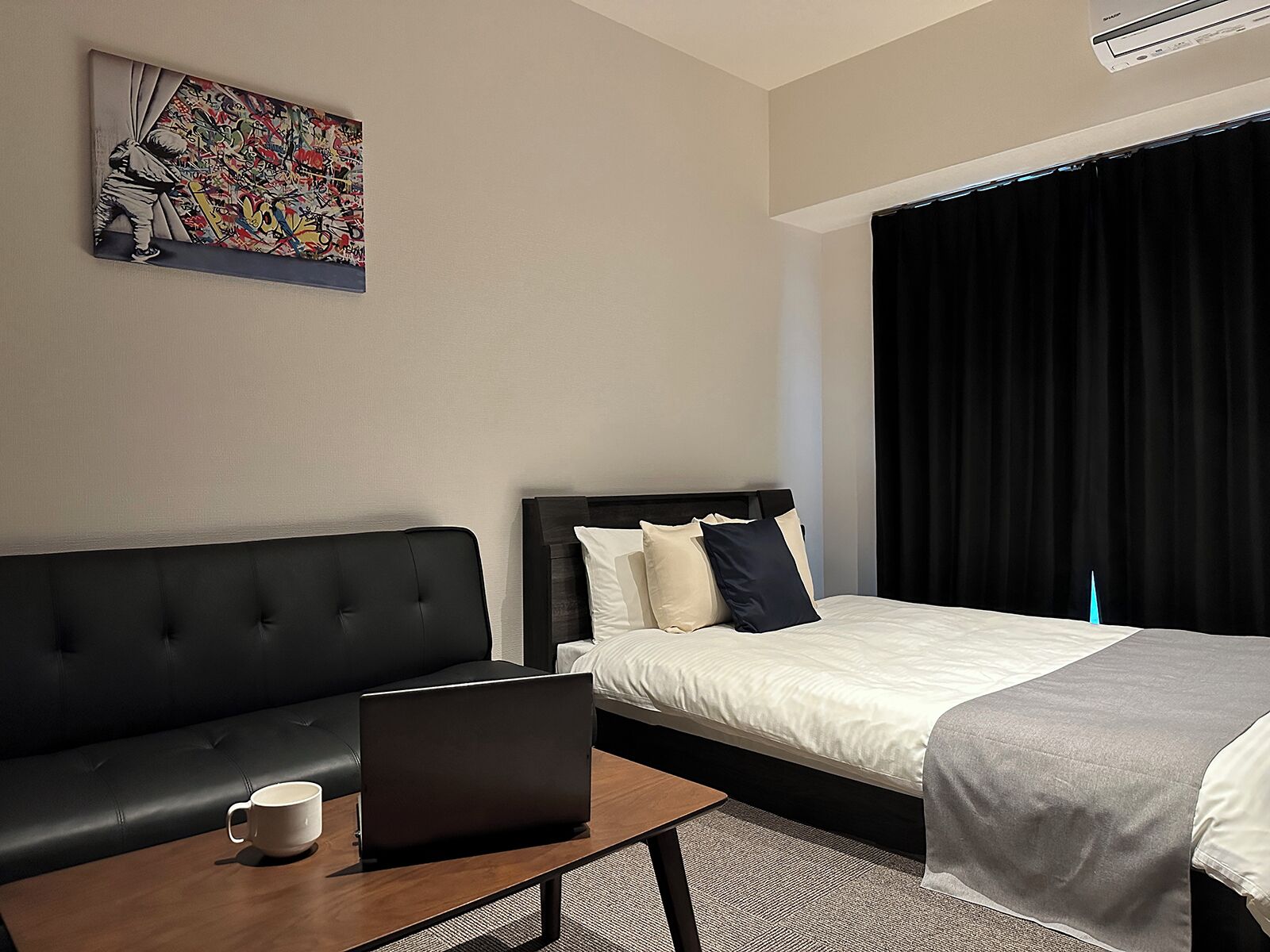 Habitación de hotel con un sofá de cuero negro, cama con almohadas blancas y azul marino, una laptop sobre una mesa de madera y un cuadro abstracto y colorido en la pared.