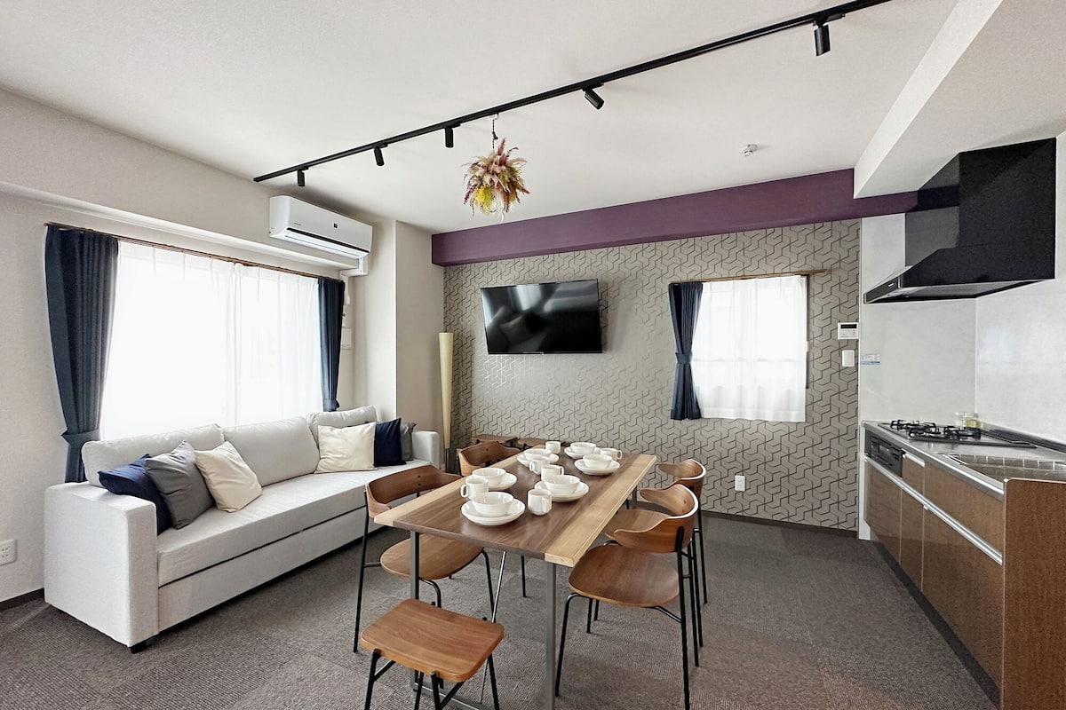 A modern living room with a sofa, dining table, and chairs. There is a flat-screen TV mounted on the wall, an air conditioning unit, and a kitchen area with a stove.