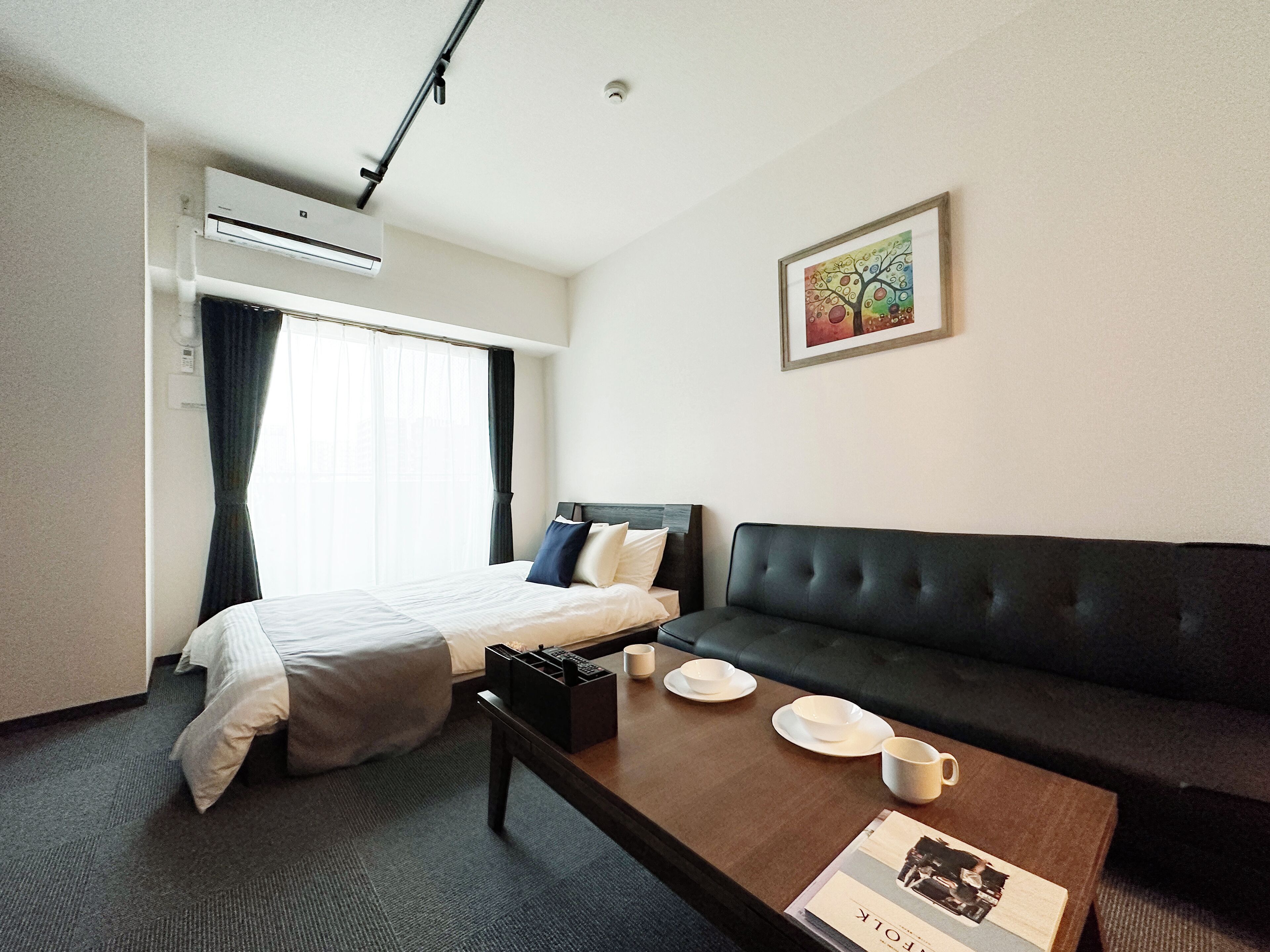 A hotel room with a bed, a black sofa, a coffee table with cups and a magazine, and a painting on the wall.