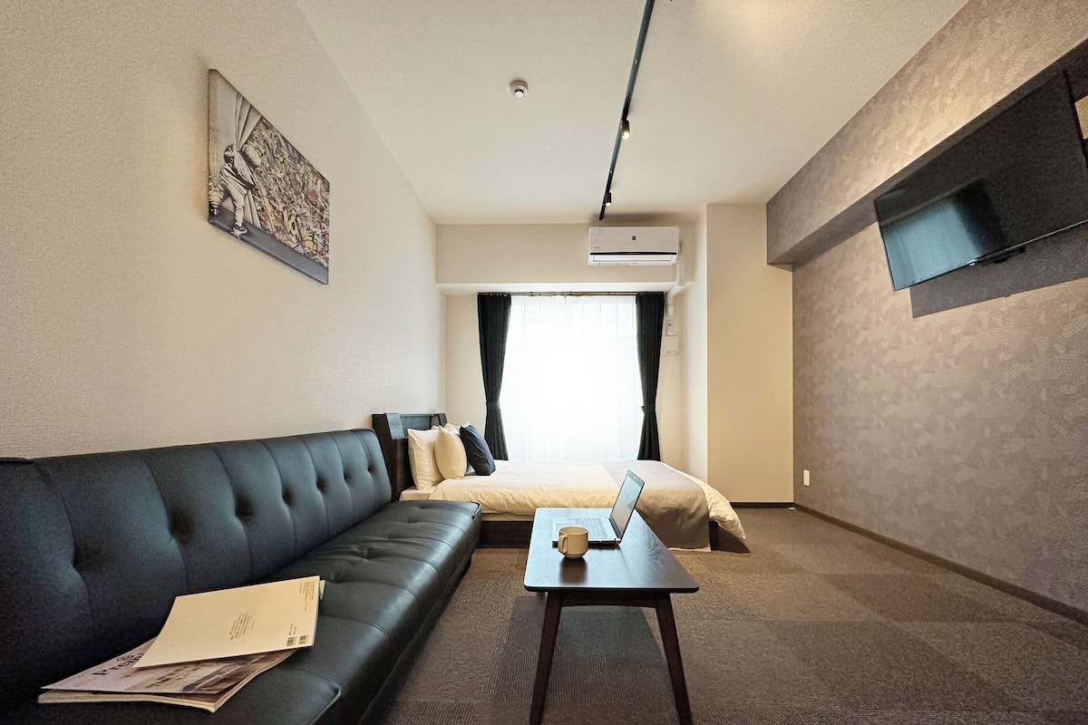 A modern hotel room with a black leather sofa, a small table with a laptop, a bed with beige bedding, and a mounted TV.