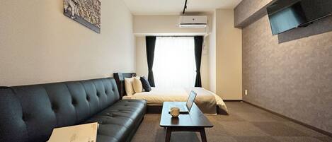 Una habitación de hotel moderna con un sofá de cuero negro, una mesita con una computadora portátil, una cama con ropa de cama color beige y un televisor de pared.