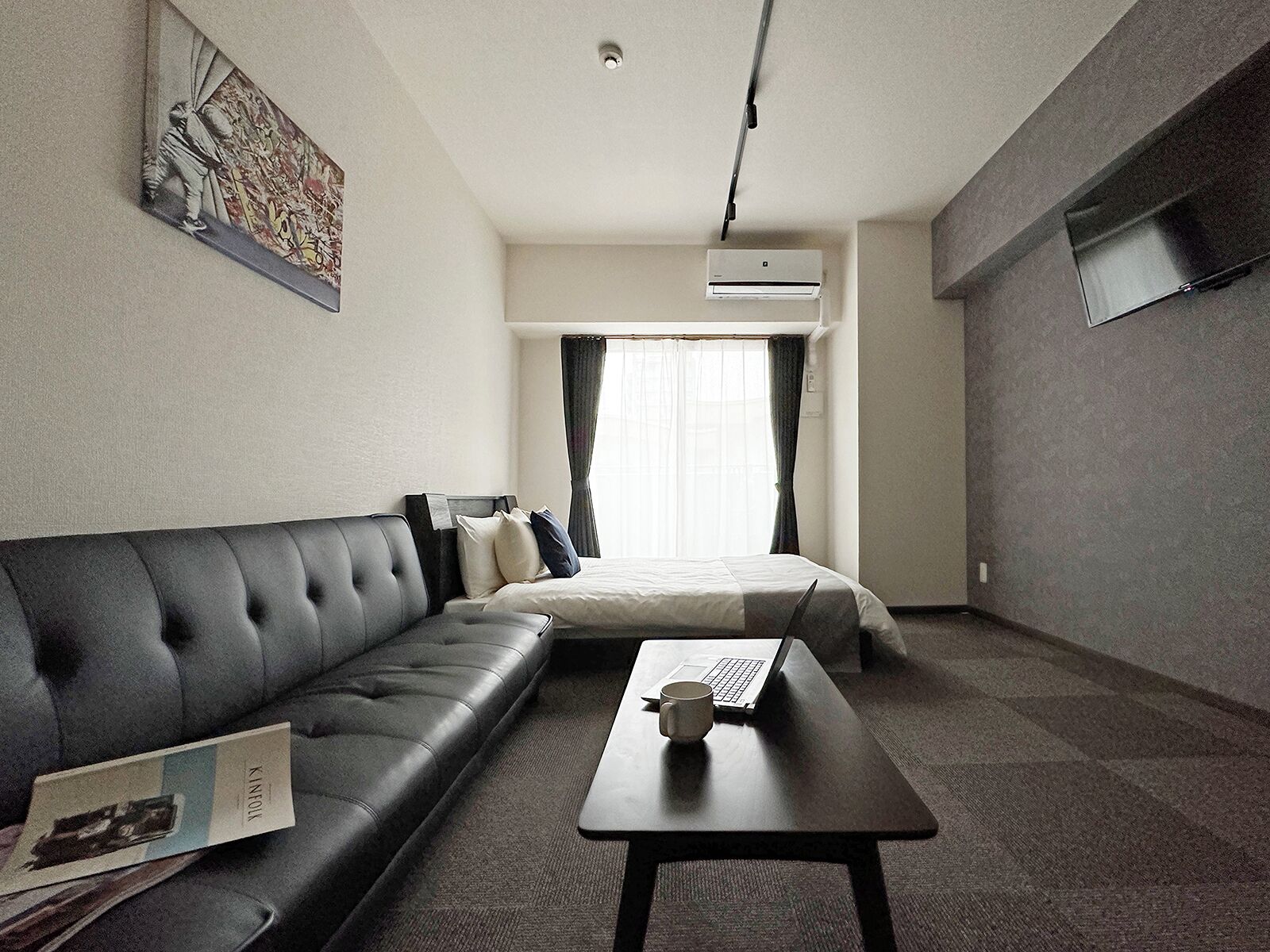 A hotel room with a black leather sofa, a single bed, a small table with a laptop and a mug, and an air conditioning unit on the wall.