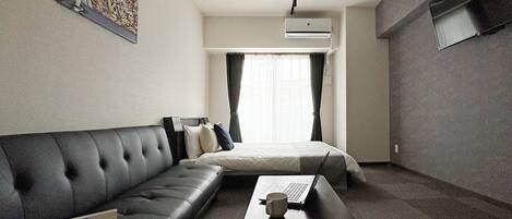 A hotel room with a black leather sofa, a single bed, a small table with a laptop and a mug, and an air conditioning unit on the wall.