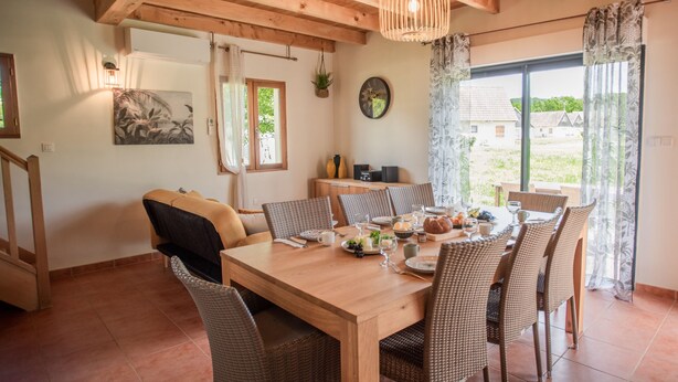 Un comedor con una mesa de madera y sillas de mimbre, una lámpara colgante y vistas al exterior a través de amplios ventanales.