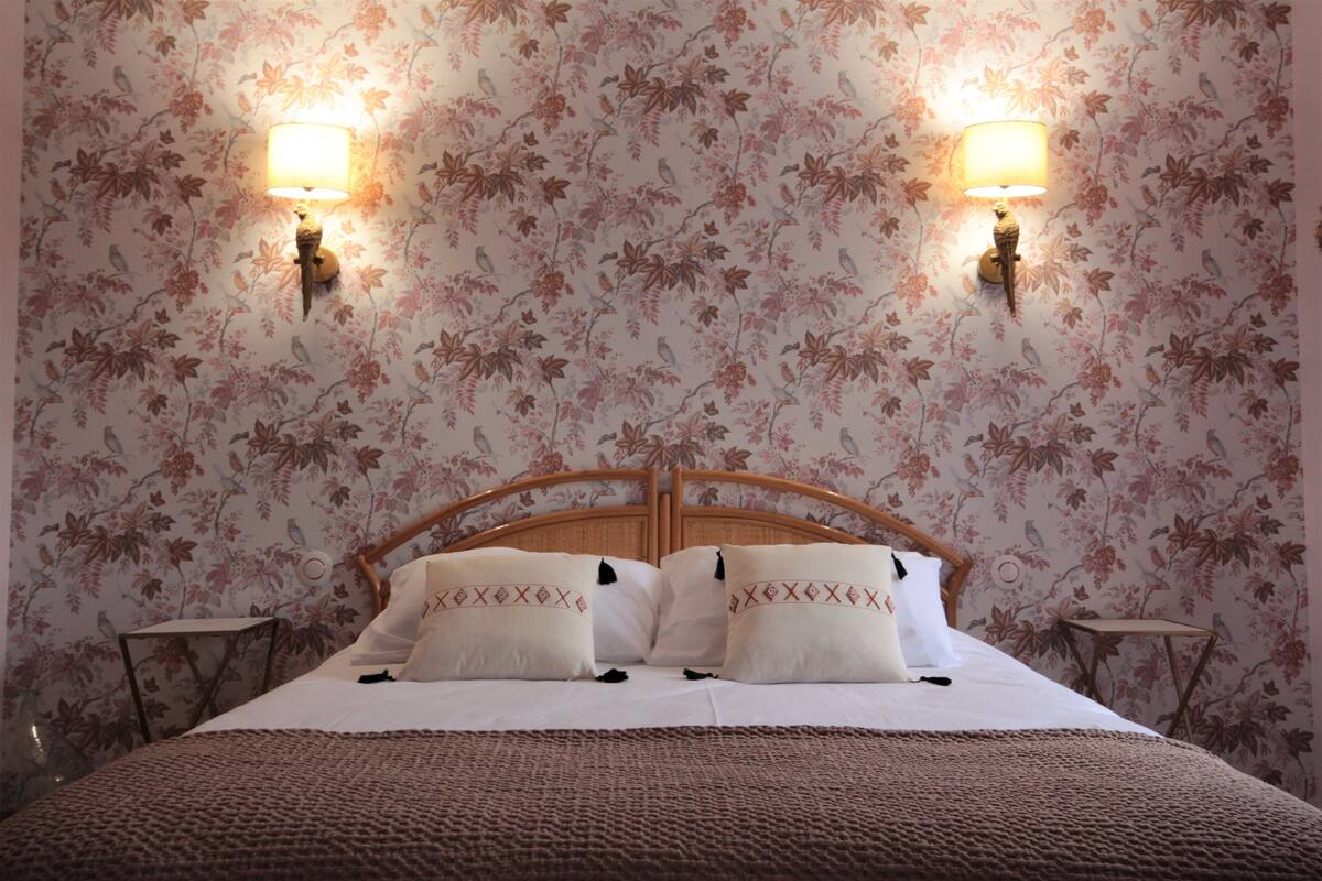 Une chambre à coucher avec un papier peint à motifs floraux, deux lampes fixées au mur, une tête de lit en bois et un lit avec une literie blanche et des coussins décoratifs.