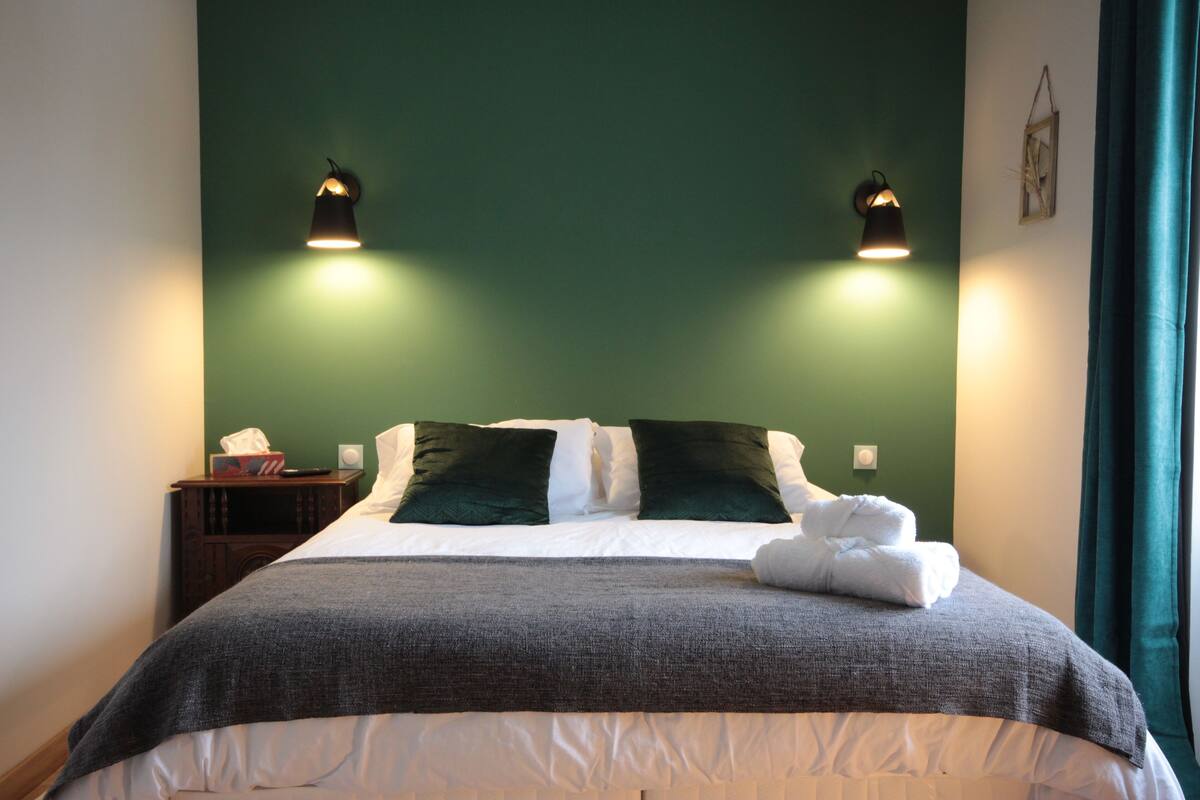 Un lit bien fait, avec une couette grise et blanche, deux oreillers vert foncé et une serviette blanche pliée posée sur le côté. Deux lampes de lecture sont fixées au mur, de part et d’autre du lit.