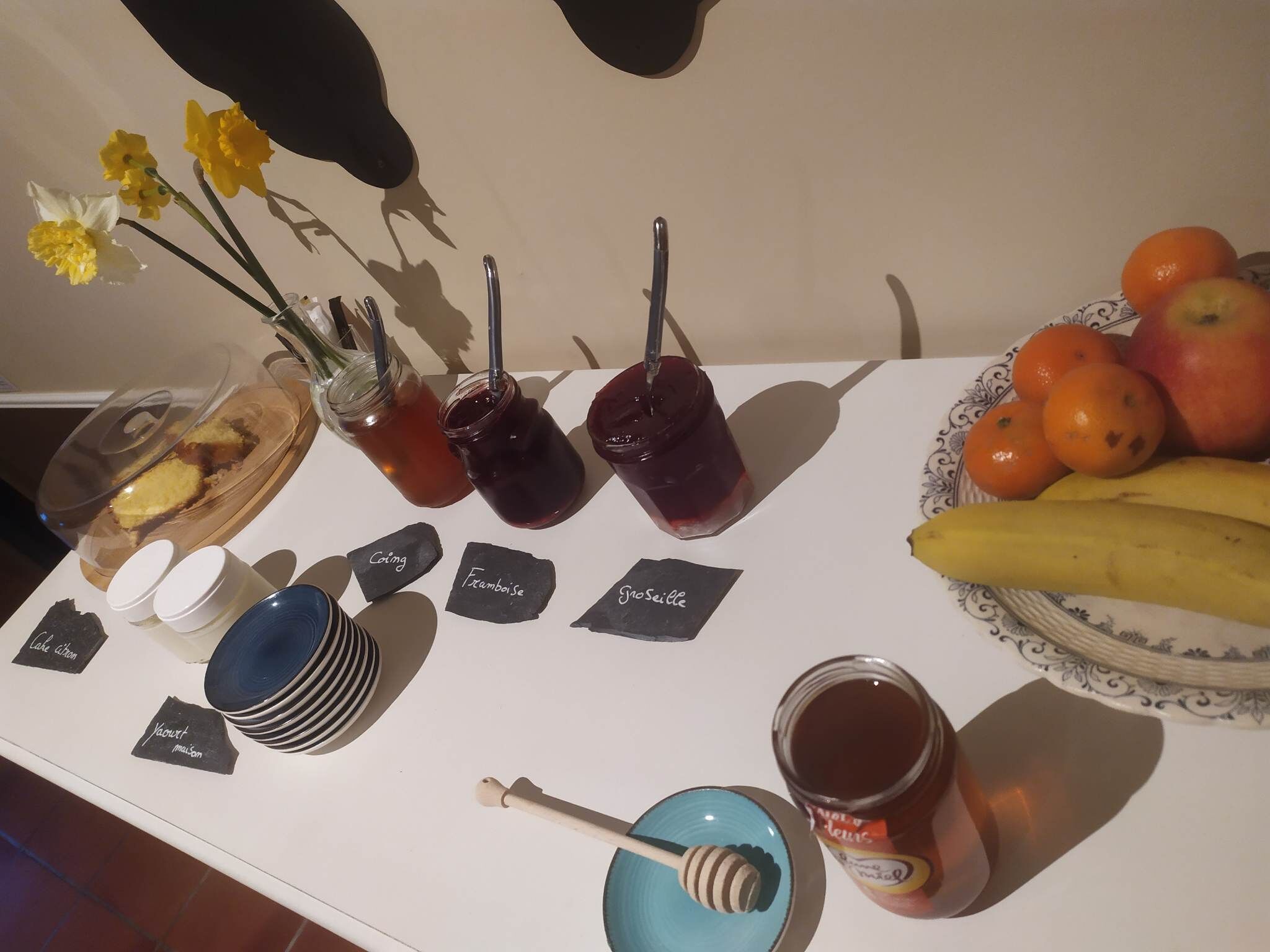 A table with various food items and condiments, including jars of preserves, a bowl of fruit, and honey.