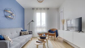 Een moderne woonkamer met een grijze bank, een klein bijzettafeltje, een tv op een witte kast, en een raam met lichte gordijnen.