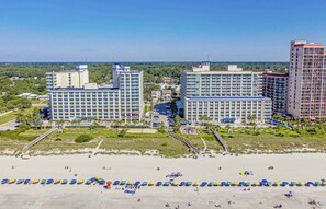 Um hotel à beira-mar com vários edifícios altos, uma praia arenosa com guarda-sóis e uma vista clara do oceano.