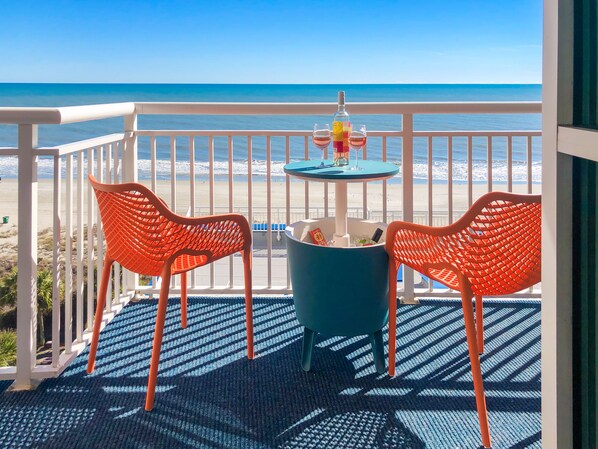 A balcony with two orange chairs, a small table with a bottle and glasses, and a view of the beach and ocean. - FUN AND SUN IN 801 Ocean Views, Resort Amenities (Myrtle Beach)