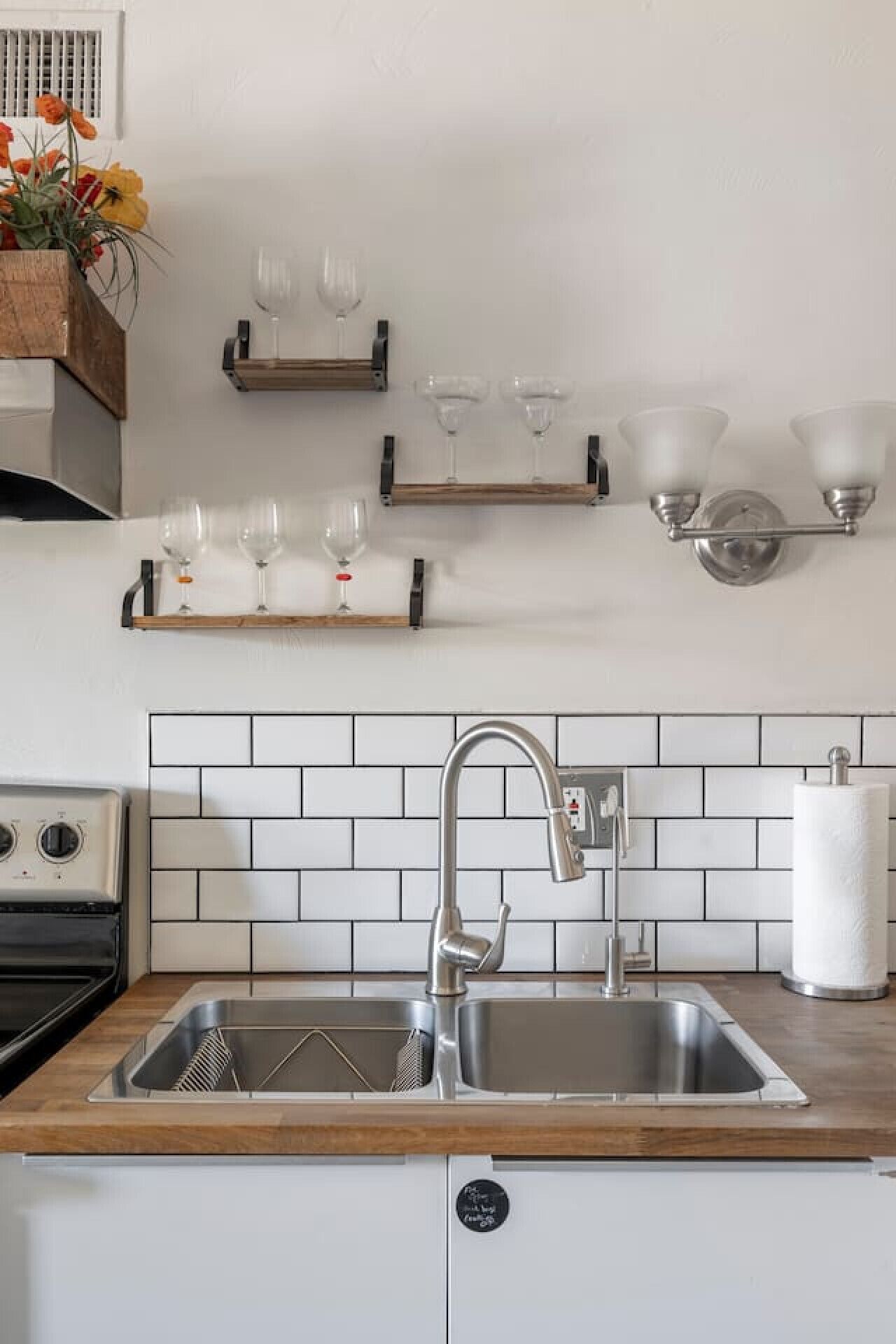 Una cocina con un lavamanos de acero inoxidable, encimera de madera, salpicadero de azulejos blancos y una lámpara fijada a la pared.