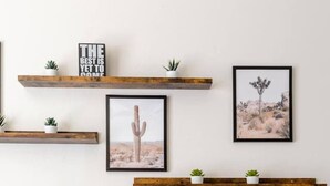 A living room with a grey sofa, three cushions, two wooden shelves with plants and framed pictures, and a cactus artwork. - Bright 4BR Getaway – Yard & Perfect Group Escape! (Tucson)