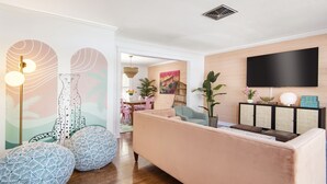 Un soggiorno moderno con un divano rosa, pouf decorati, una TV a schermo piatto e, sullo sfondo, una zona pranzo.