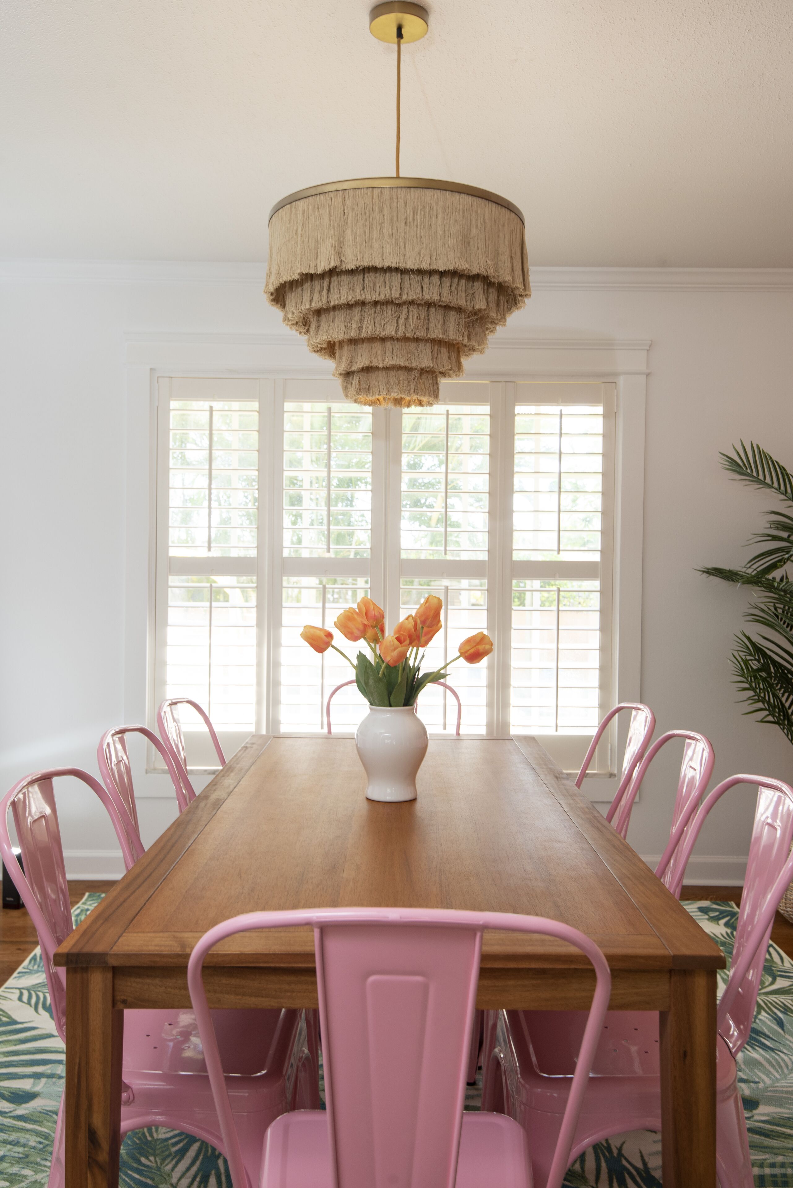 A dining room with a wooden table, pink chairs, a white vase with orange flowers, and a large window with white shutters.