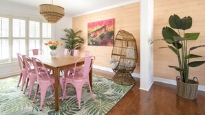 Zona pranzo con tavolo in legno, sedie rosa, tappeto verde decorato, lampadario a sospensione, quadro appeso al muro e sedia in vimini.