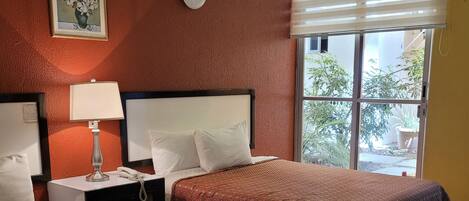 Una habitación de hotel con cama, mesita de noche con lámpara, un reloj y una ventana con persianas.
