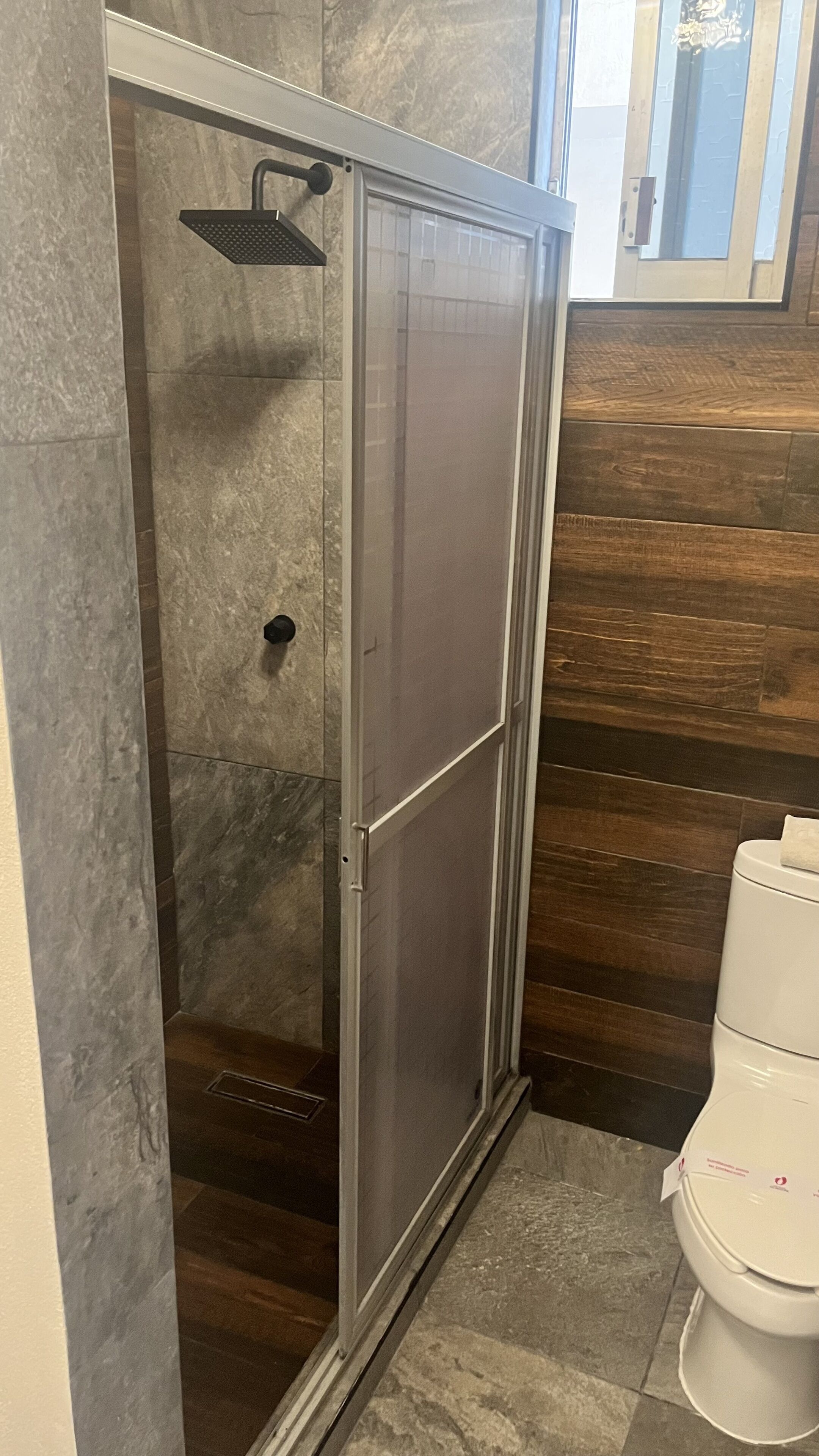 Una ducha a nivel con puerta de cristal, un cabezal de ducha y un inodoro en un baño con azulejos de mármol y madera.