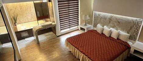 A modern bedroom with a large bed, a bedside table, a nightstand, a window with blinds, and a bathroom with a sink and mirror.