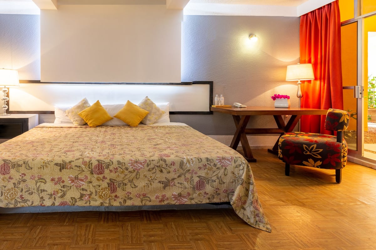 A hotel room with a bed, bedside lamps, a small table, a chair, and a view of the outside.
