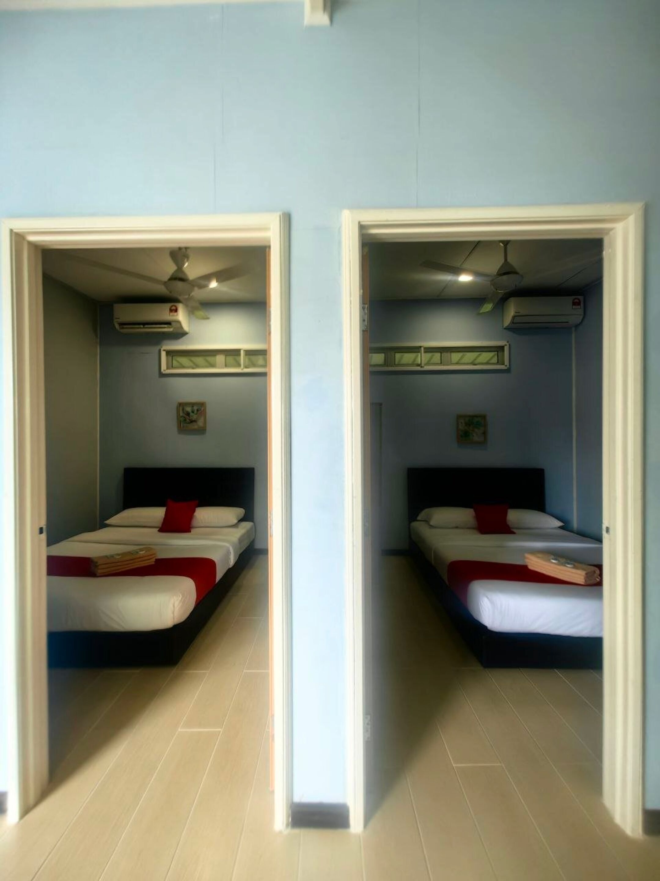 Habitación de hotel con dos camas, cada una con almohada roja y ropa de cama blanca. Se observan ventiladores de techo y unidades de aire acondicionado sobre las camas.