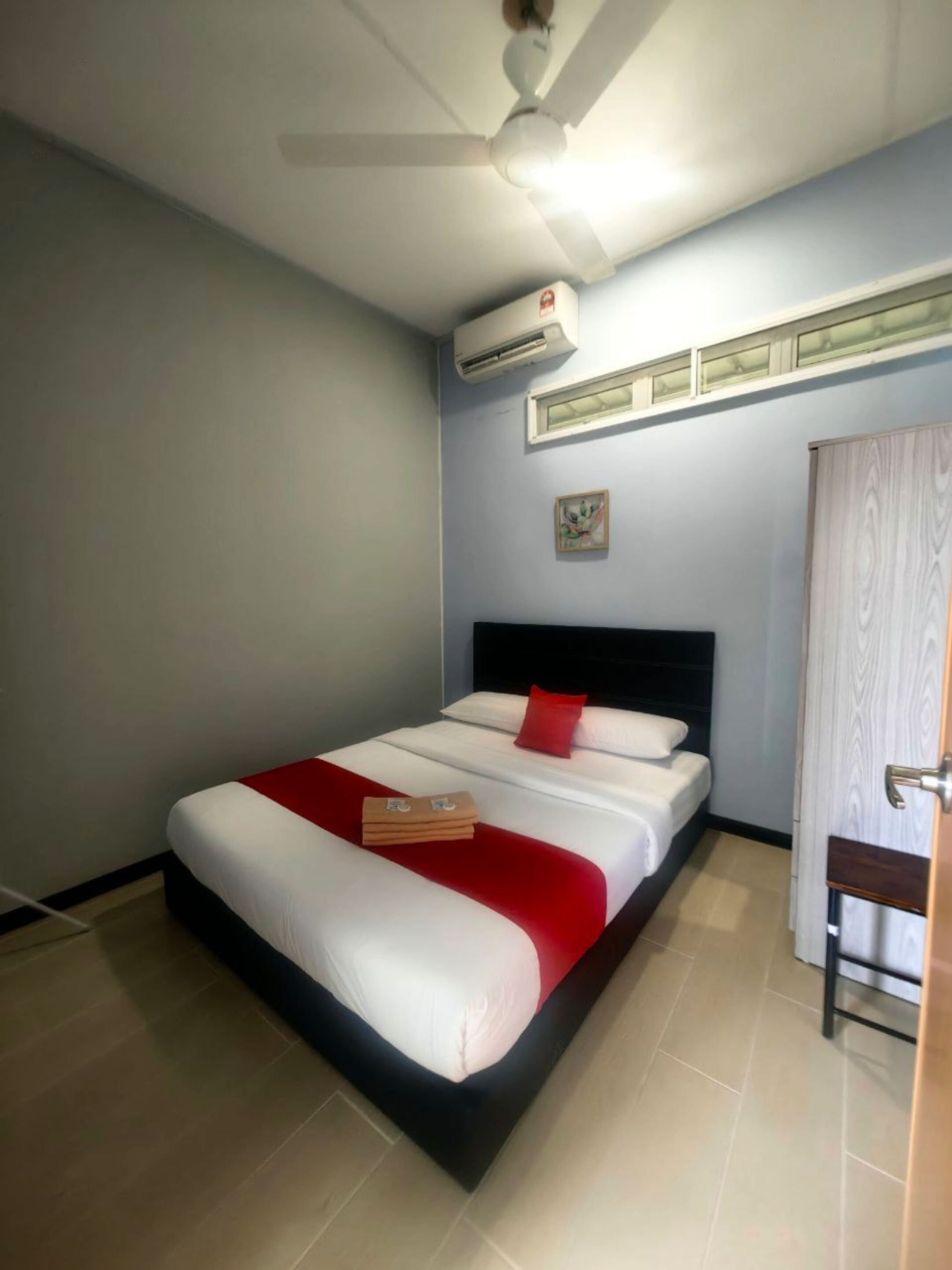 A hotel room with a bed, a ceiling fan, an air conditioning unit, a window, and a door.