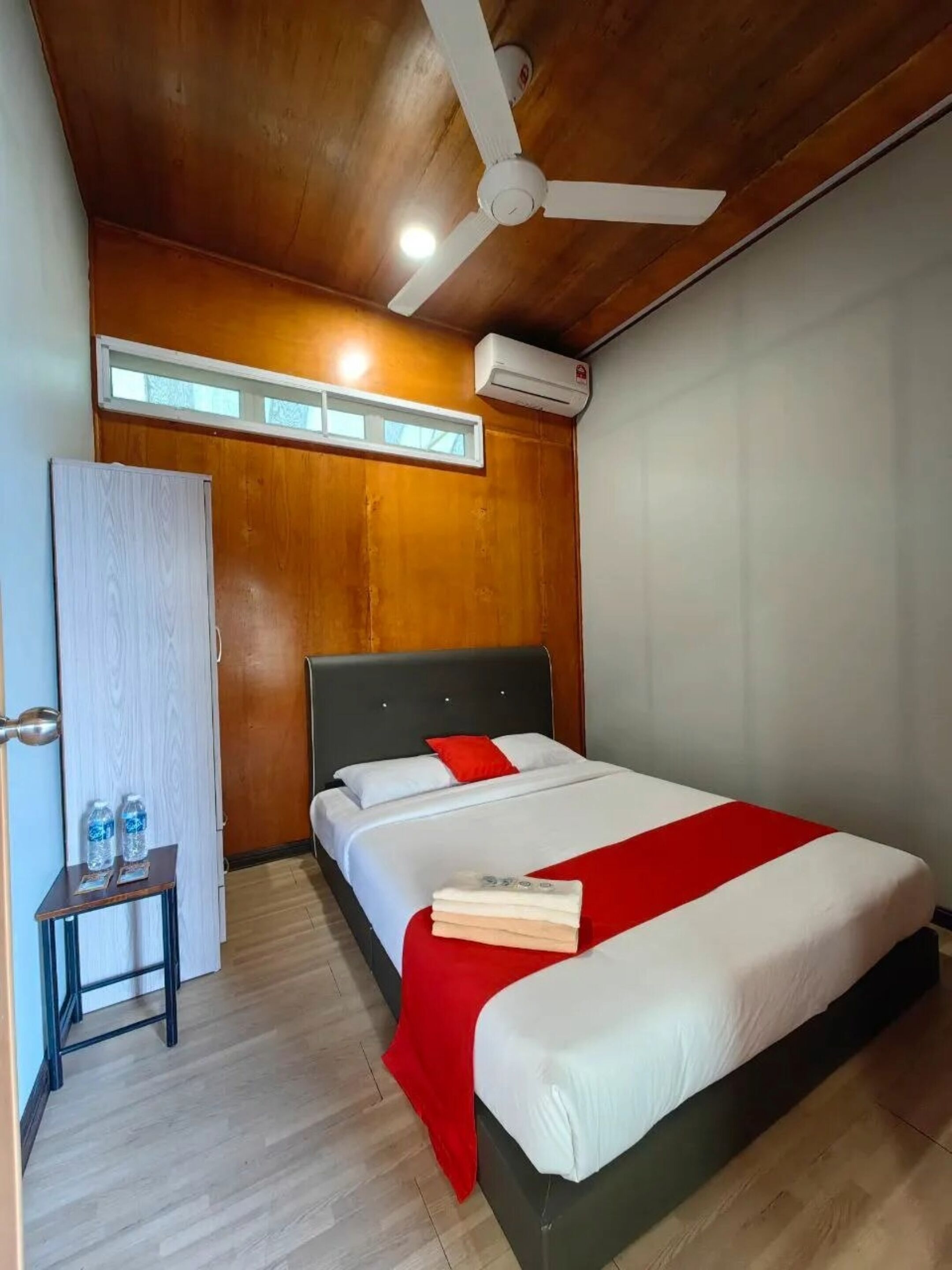 Quarto de hotel com teto de madeira, ventilador de teto, ar condicionado, cama com roupa de cama branca e detalhes vermelhos, mesa de cabeceira com garrafas de água e janela com vista.