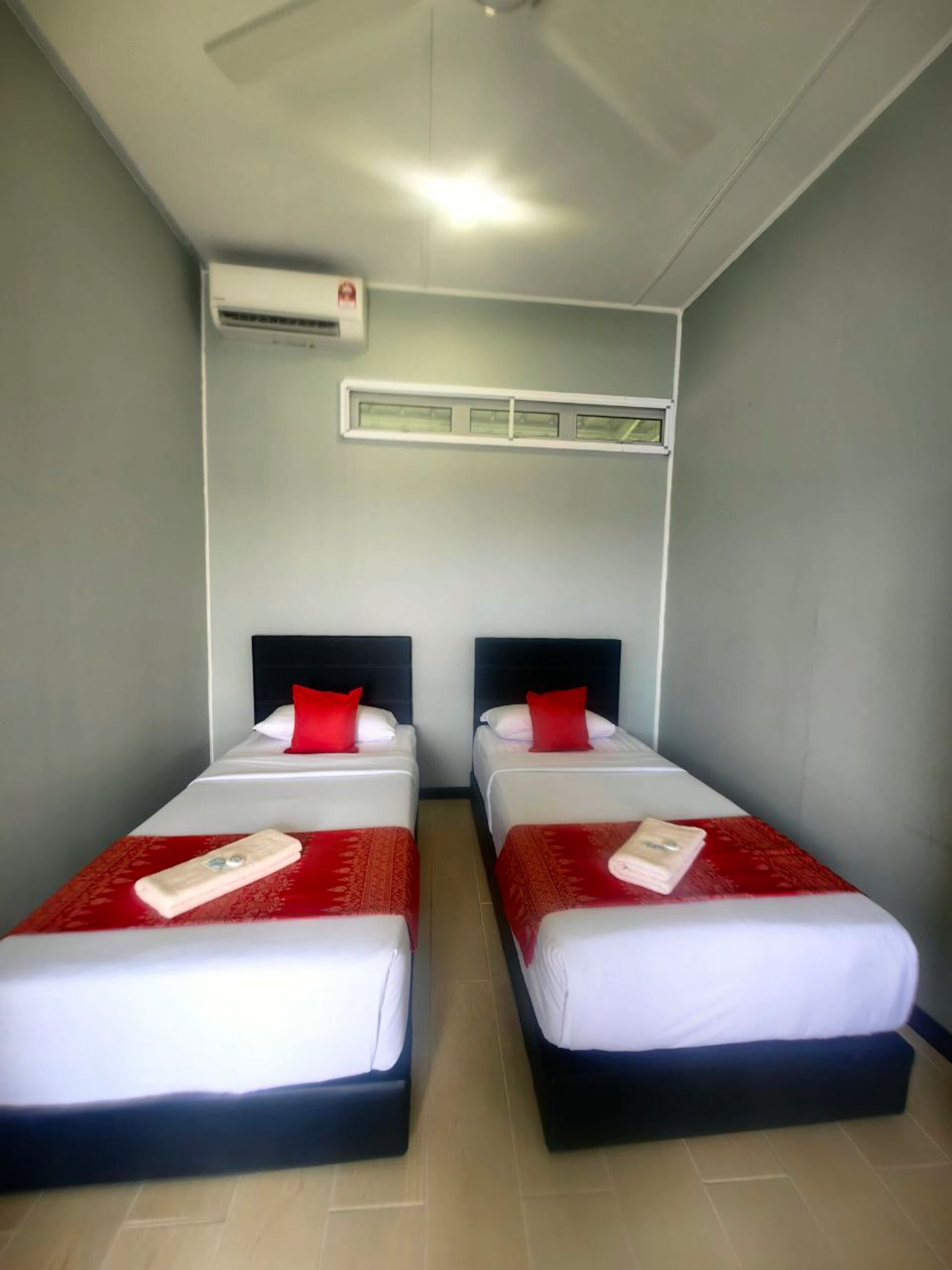 A room with two beds, each with a red and white bedspread, and a white air conditioning unit above.